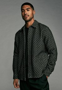 Black and white patterned shirt with a diamond design, featuring a button-up front, collar, and relaxed fit. Worn over a black top.