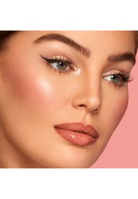 Primo piano di un volto con una pelle liscia, zigomi ben definiti, labbra nude e lucide, eyeliner a forma di ala e ciglia morbide su uno sfondo rosa.