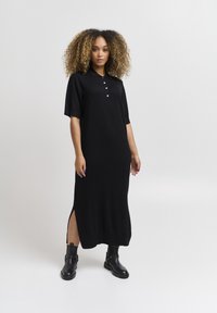 Vestito nero di lunghezza al ginocchio con maniche corte, colletto polo, tre bottoni e spacchi laterali. Tessuto testurizzato. Indossato con stivali neri alla caviglia.