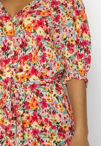 Abito floreale con un motivo multicolore di fiori rossi, rosa e gialli. Tessuto leggero, scollo a V, chiusura con bottoni e vita regolabile con laccio.