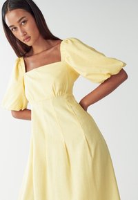 Žlté šaty s puffovanými rukávmi, štvorcovým výstrihom, zhromaždenými detailmi a plisovanou sukňou. Vyrobené z ľahkej tkaniny s jemnou textúrou.