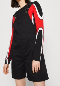 Svarta jeanshängselbyxor med silverfästen, som har röda långärmade tröjor med vita och svarta ränder på armarna, vilket fulländar en sportig design.