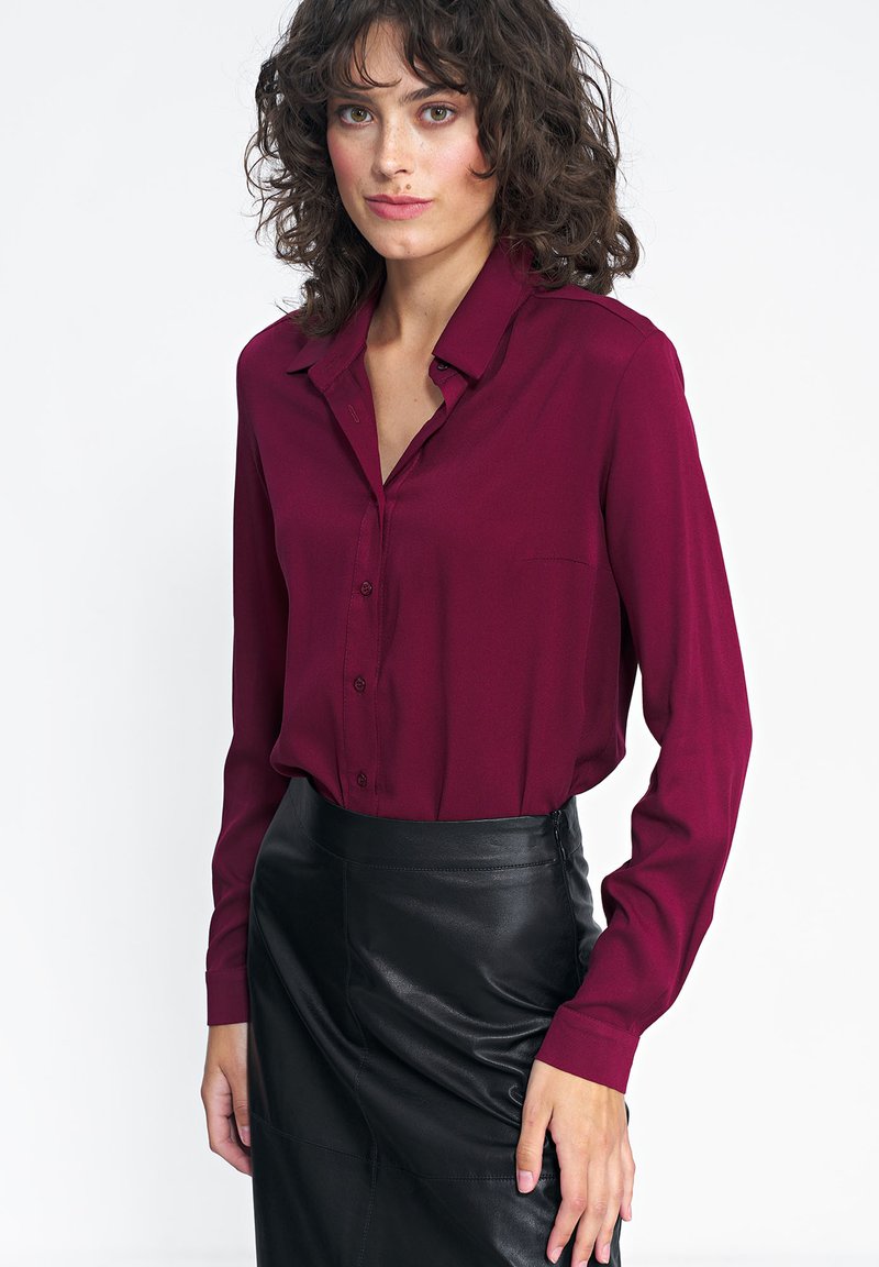 Bordowa bluzka z długim rękawem, kołnierzykiem i guzikami, z kieszonką na piersi oraz gładkiego materiału, zestawiona z czarną skórzaną spódnicą.