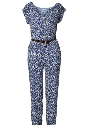 Frau trägt einen blau-weiß gemusterten Overall mit schwarzem Gürtel, einen grau gestrickten Cardigan über die Schultern gelegt und weiße Schnürschuhe.