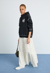 Svart hettegenser med frontlomme og hvit logo, kombinert med et hvitt lagdelt maxiskjørt med rutemønster, og hvite slip-on sko.