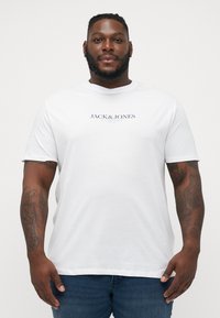 Bijela majica kratkih rukava od pamuka, s otisnutim logom "JACK & JONES" u mornarsko plavoj boji preko prsnog dijela. Ležeran kroj.