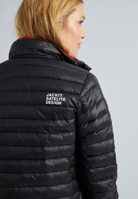 Veste en duvet noire avec un design matelassé, dotée d'un col montant et d'accents rouges, incluant le logo "JACK1T SATELITE DESIGN" au dos.