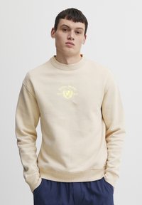 Sudadera beige con cuello redondo, puños y dobladillo de canalé. Presenta un diseño de logo amarillo en el pecho. Fabricada en un tejido suave. Corte casual.