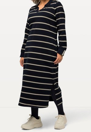 Femme portant une longue robe bleu marine à rayures horizontales blanches, fente latérale, leggings noirs et baskets blanches.