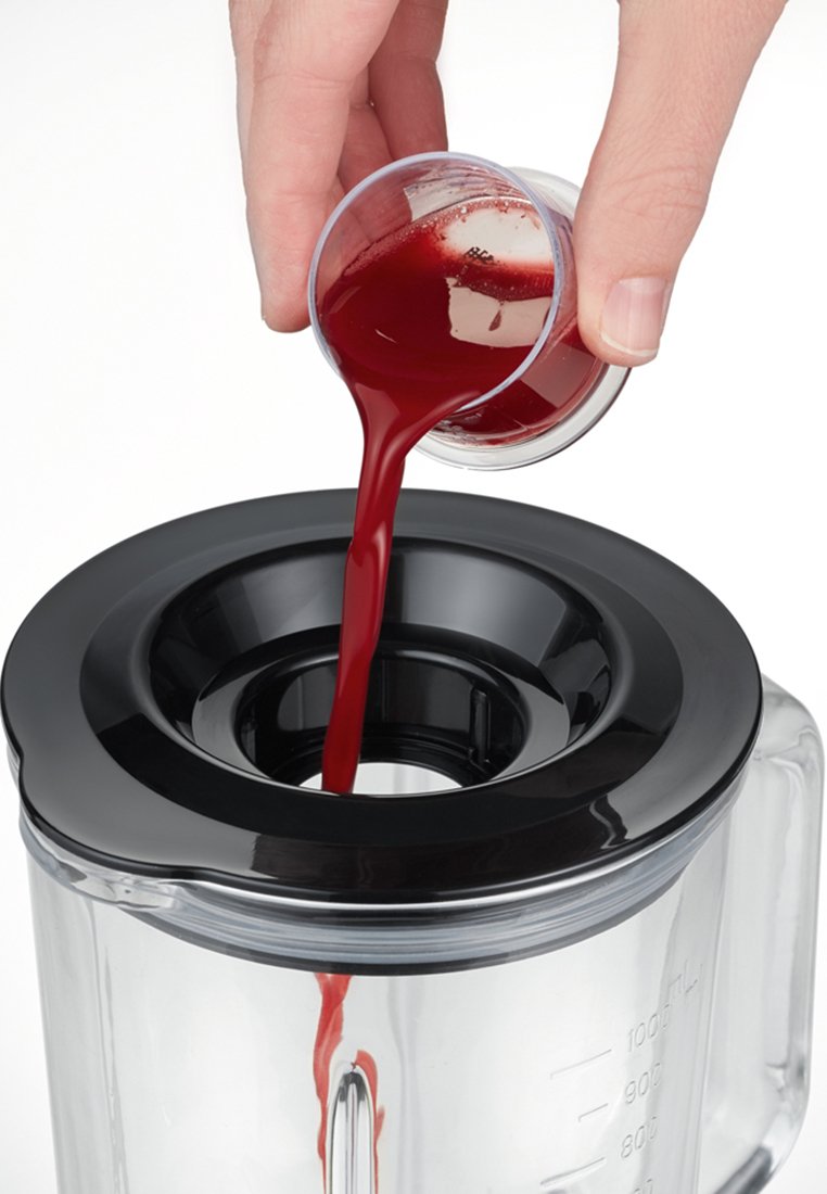 Eine Hand, die roten Flüssigkeit aus einem kleinen durchsichtigen Becher in einen Mixer mit schwarzem Deckel auf weißem Hintergrund gießt.