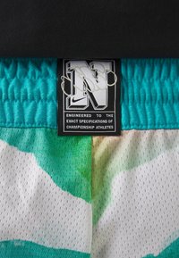 Étiquette de vêtement noire avec une lettre blanche N et le logo Nike, cousue sur un tissu mesh sportif bleu-vert à motif. Le texte indique "Engineered to championship athletes."