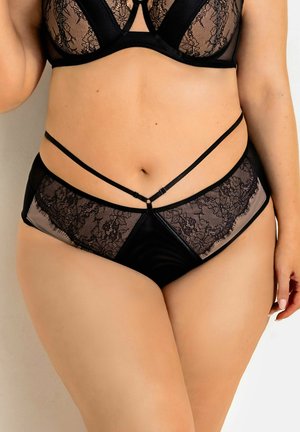 Kobieta ubrana w czarny koronkowy zestaw bielizny z przezroczystymi panelami i ozdobnymi cienkimi paskami wokół talii, pozująca na jednolitym tle.