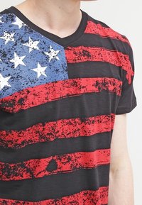 Svart T-shirt med en sliten design av röda och svarta horisontella ränder, med en blå sektion och vita stjärnor som påminner om en flagga.