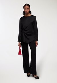 Top lungo a maniche nere in satin con dettaglio attorcigliato, abbinato a pantaloni neri. Gli accessori includono una piccola borsa rossa e scarpe con tacco nere.