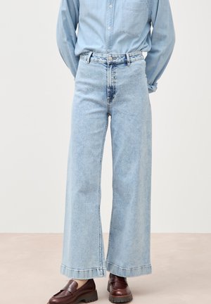 Person trägt hellblaue Jeans mit weitem Bein, ein helles Jeanshemd und dunkelbraune klobige Loafers und steht vor einem einfarbigen Hintergrund.
