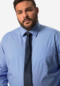 Světle modrá, dlouhá košile s knoflíky a špičatým límečkem a kapsou, doplněná tmavě námořnickou kravatou. Hladká textura látky.