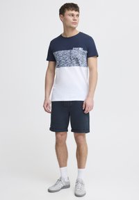Marineblå og hvit fargeblokert t-skjorte med en teksturert øverste stripe og en brystlomme, sammen med marineblå shorts og lysegrå sneakers.