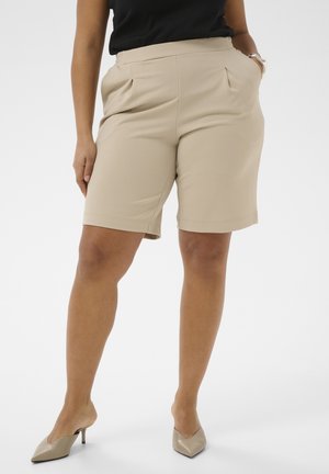 Vrouw draagt beige, op maat gemaakte shorts tot net boven de knie, een zwart topje en beige pumps met een spits toelopende neus, staat met één hand in de zak tegen een witte achtergrond.