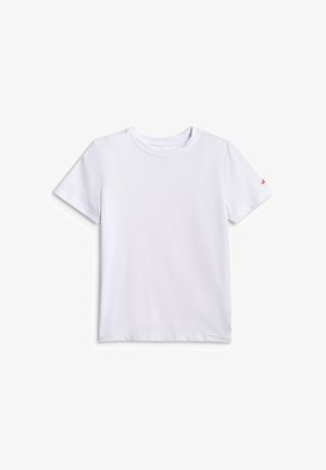 Enfant blond debout, portant un t-shirt blanc, un short noir, des chaussettes blanches à rayures noires et des baskets noir et blanc, sur un fond uni.