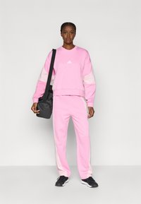 Rozā džemperis ar baltām detaļām, atbilstošas rozā sporta bikses un melnas sporta kurpes. Plecu soma ir pārklāta pāri plecam.