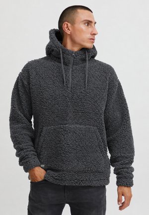 Homme portant un sweat à capuche en polaire texturée gris foncé avec poche avant, debout avec une main dans la poche, regardant sur le côté.