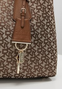 Sac à main DKNY marron avec motif répétitif de logo, étiquette en cuir embossée du nom de la marque, et porte-clés doré attaché avec une clé et un charme de marque.