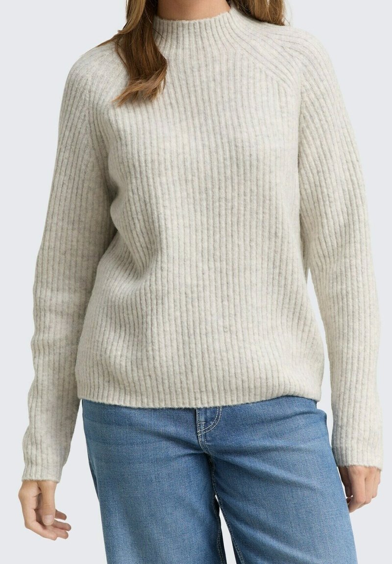 Femme portant un pull côtelé beige clair et un jean bleu, debout devant un fond uni.