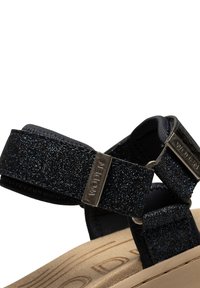 Mustikka ja sininen sandaali, jossa on kimaltavat hihnat, säädettävät suljettavat osat ja vaaleanbeige pehmustettu pohja, jossa on kohotettu koristelu.