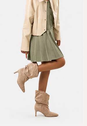 Femme portant des bottes beige à talon boudinées avec une jupe plissée verte et une veste courte beige, posant avec une jambe levée.
