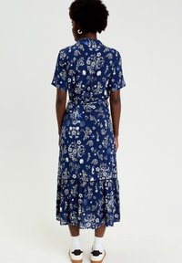 Mujer de espaldas vistiendo un vestido midi azul con flores, mangas cortas, dobladillo con volantes, calcetines blancos y zapatillas blancas.