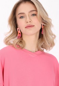 Camisa de gran tamaño en color rosa con cuello en V, de tejido texturizado. Pendientes geométricos grandes en rosa, naranja y rosa claro. Diseño simple y moderno.