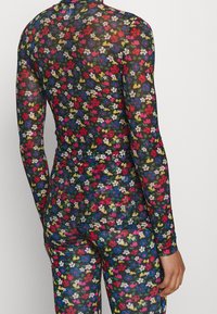 Person trägt ein eng anliegendes schwarzes blickdichtes Outfit aus transparentem Stoff mit kleinem, buntem Blumenmuster, von hinten vor einem schlichten Hintergrund gezeigt.