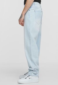 Light blue denim jeans with a relaxed fit, five-pocket design, and contrasted stitching. Worn with white sneakers featuring black accents.