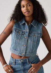 Gilet en denim bleu clair, court, avec une fermeture à boutons, deux poches poitrine et un col classique. Présente un ourlet effiloché et des détails de couture subtils.