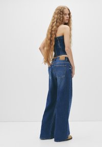 Outfit in denim composto da un top corto senza spalline e jeans a gamba larga, entrambi in una tonalità blu medio, con dettagli di borchie sottili lungo il lato.