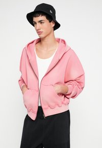 Jong persoon draagt een losse roze hoodie met rits over een wit tanktopje, zwarte broek en een zwarte bucket hat, staat met handen in de zakken.