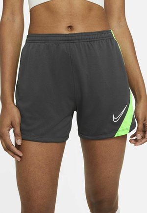 Osoba ubrana w ciemnoszare sportowe szorty Nike z neonowozielonymi akcentami i biały stanik sportowy, stoi z ramionami luźno opuszczonymi wzdłuż boków.