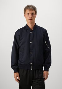 Man wearing a navy bomber jacket with snap buttons and zippered pocket on the sleeve, standing against a plain light background.
