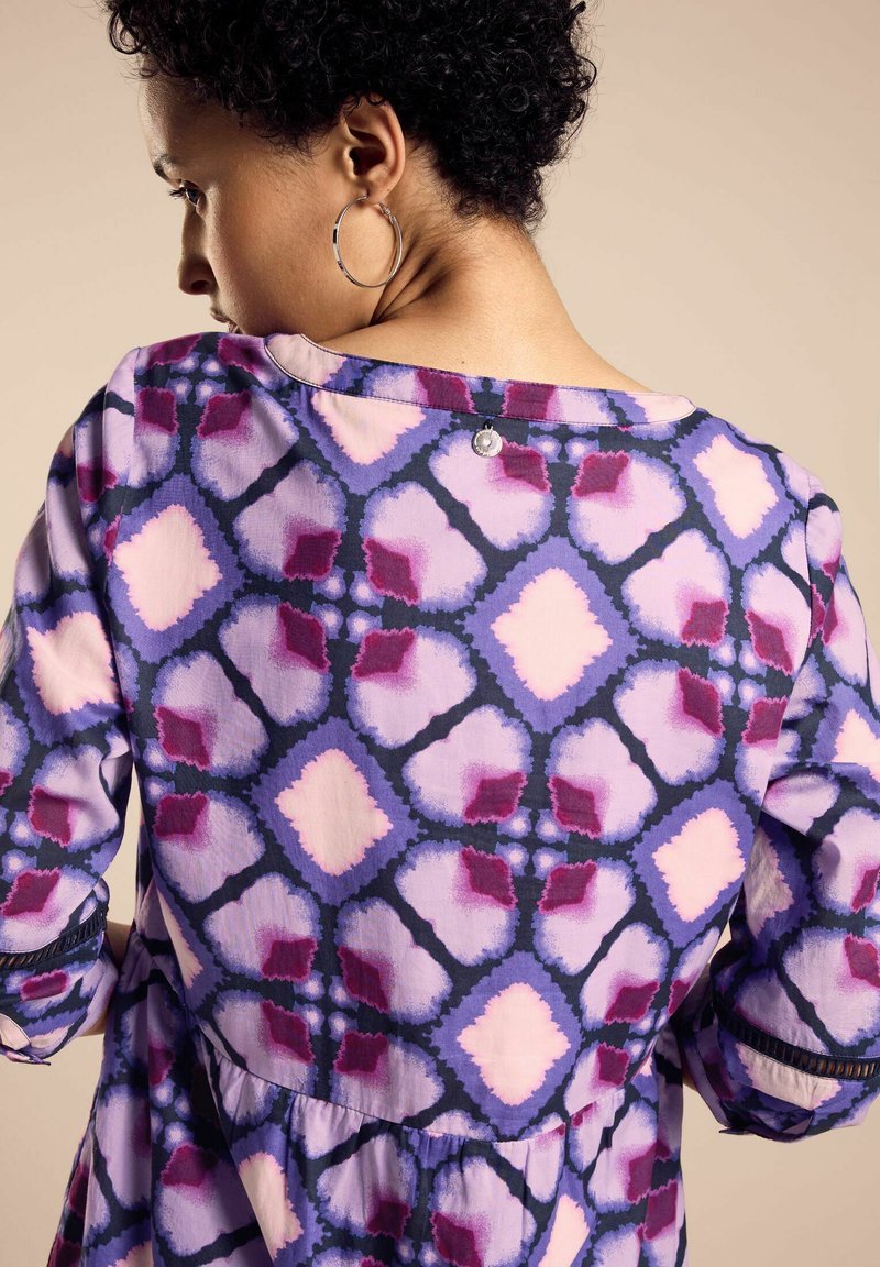 Lila gemusterte Bluse mit lockerer Passform, die eine Mischung aus Rauten in Pink und Dunkelblau zeigt, sowie einem Knopfdetail am hinteren Halsausschnitt.