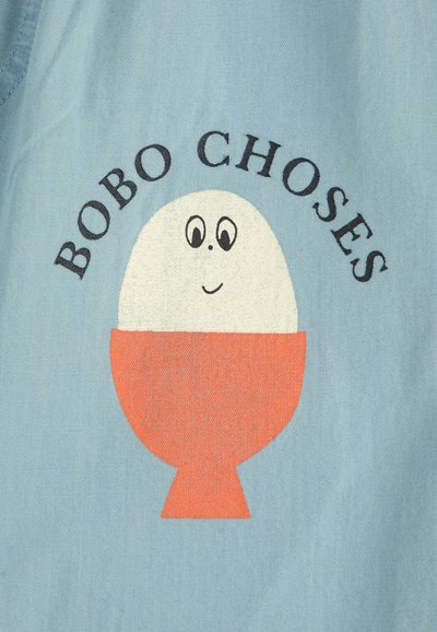 Svetlomodrá látka s hravým motívom usmievajúceho sa vajíčka v oranžovej miske. Text "BOBO CHOSES" sa klenie nad dizajnom.