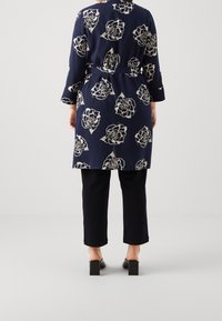 Marinblå blommig kappa med bälte, krämfärgade rosor, långa ärmar och rak skärning, kombinerad med svarta korta byxor och skor med fyrkantig klack.