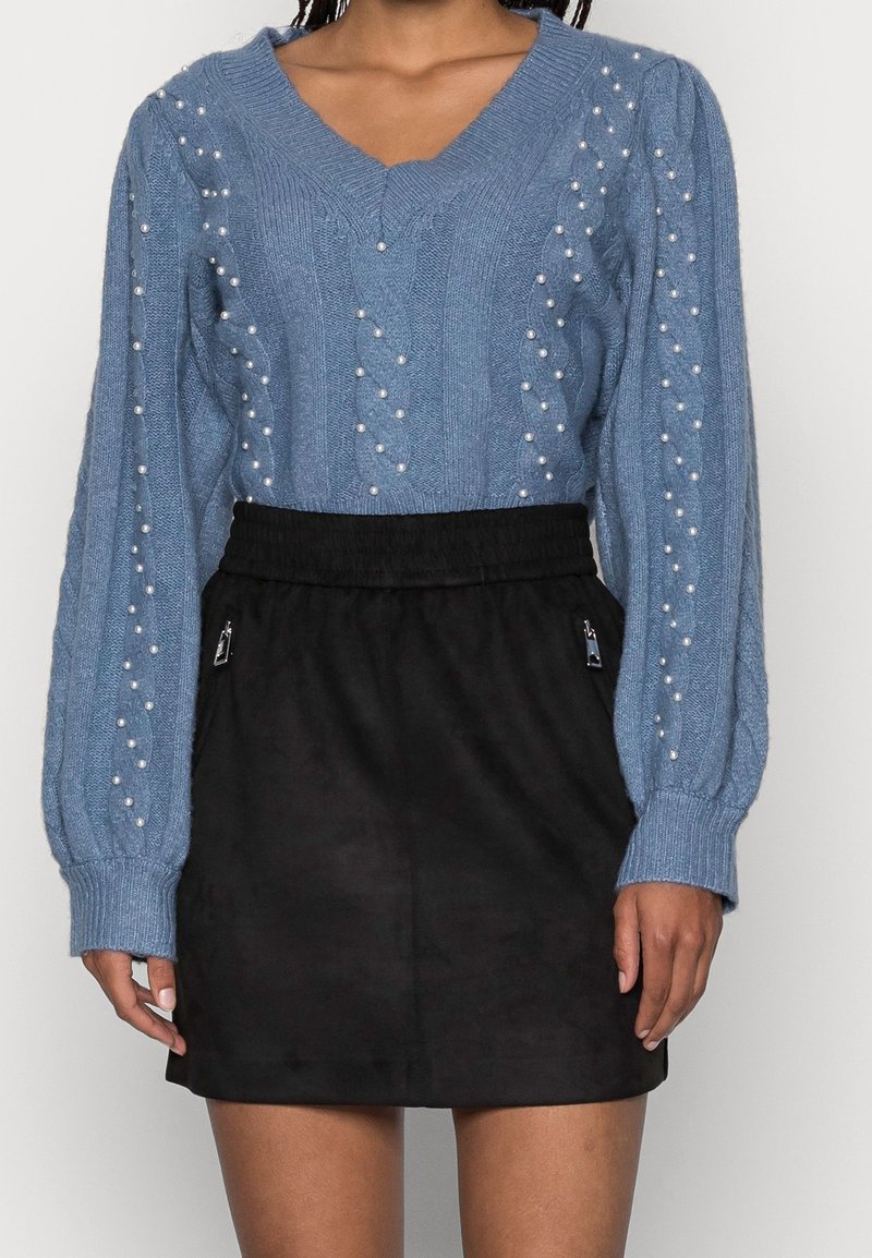 Pull en maille bleu orné de perles, associé à une jupe noire taille haute avec poches zippées.