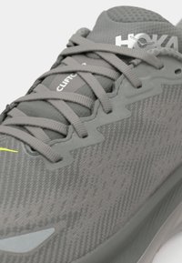 Scarpa da corsa grigia con tomaia in mesh testurizzato, accentuata da lacci piatti e un dettaglio giallo. Presenta un logo in rilievo e un design della suola flessibile.
