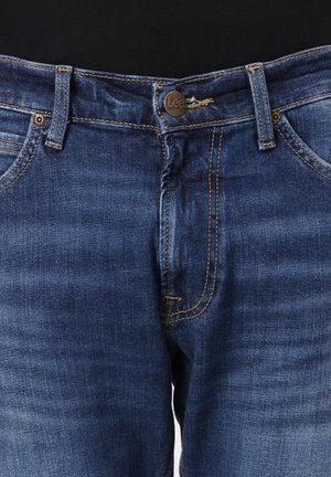 Jean en denim bleu avec fermeture à boutons devant, passants pour ceinture, rivets en cuivre et coutures orange.