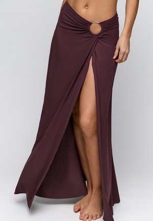 Vrouw draagt een bordeauxrode maxirok met een hoge split tot op het bovenbeen, vastgehouden door een cirkelvormige ringdetail in de taille, staat blootsvoets op een grijze achtergrond.