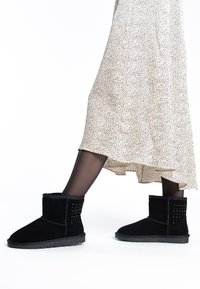 Botas de tobillo de ante negro con forro de pelaje sintético, que presentan acentos de tachuelas en el costado y una suela de goma texturizada para mayor agarre.