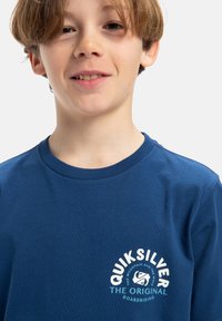 Tmavě modré bavlněné tričko s bílým a tyrkysovým potiskem loga na levé přední straně, s klasickým kulatým výstřihem a krátkými rukávy.