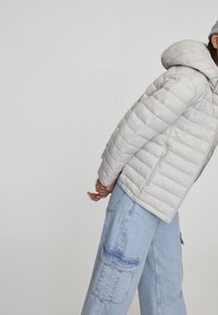 Personne portant une doudoune gris clair et un jean cargo bleu clair, debout de profil les mains jointes dans le dos sur un fond uni.