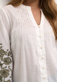 Top bianco in lino con pattina, caratterizzato da ricami testurizzati sulle maniche e bottoni rotondi decorativi lungo la parte anteriore.