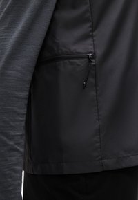 Μαύρο ελαφρύ μπουφάν με ομαλή εξωτερική επιφάνεια, που διαθέτει μια τσέπη με φερμουάρ στο πλάι και αντίθετο γκρι υφαντό ύφασμα στο μανίκι.